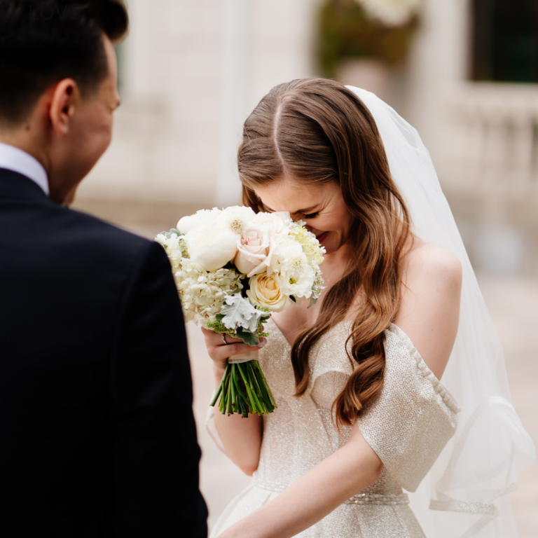 Bride at wedding venue in Dallas laughing into her bouquet with groom watching.